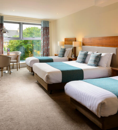 Bright hotel room with three beds, teal accents, large window, and seating area.