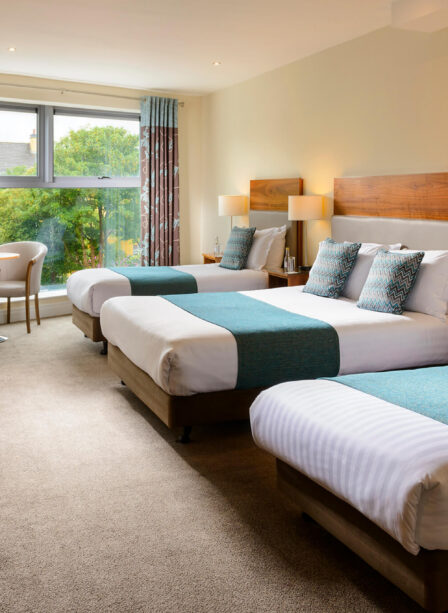 Bright hotel room with three beds, teal accents, large window, and seating area.