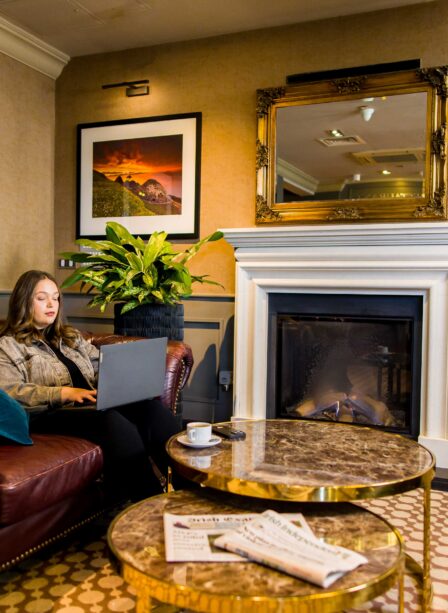 Woman relaxing on a leather sofa with a laptop by a cosy fireplace at Scotts Hotel Killarney.
