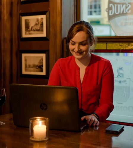 Woman in red shirt smiles while working on laptop at cosy hotel bar with candle and wine.