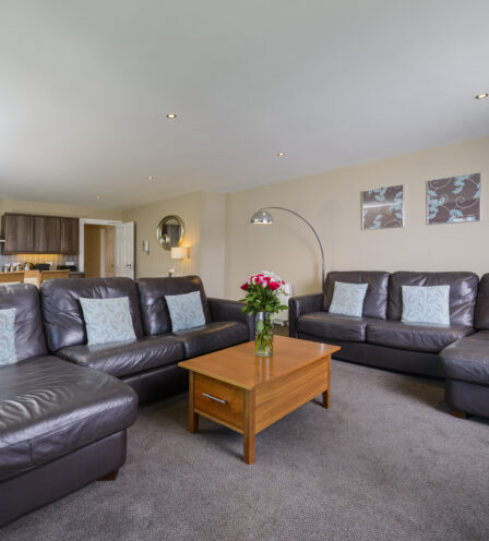 Spacious hotel lounge with cosy leather sofas, floral decor, and modern kitchen area.
