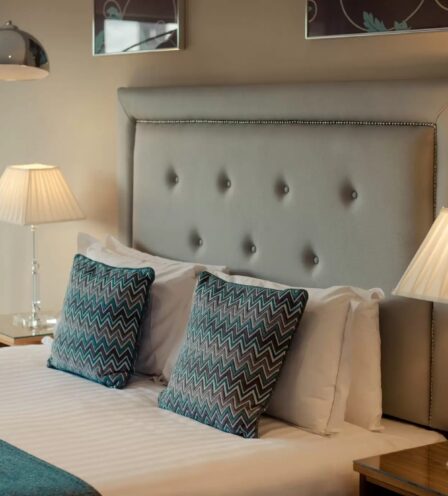 Elegant hotel room with a plush bed, two teal zigzag cushions, and soothing warm lighting.
