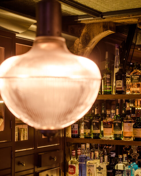 Cosy hotel bar with glowing light fixture and shelves filled with various spirits.
