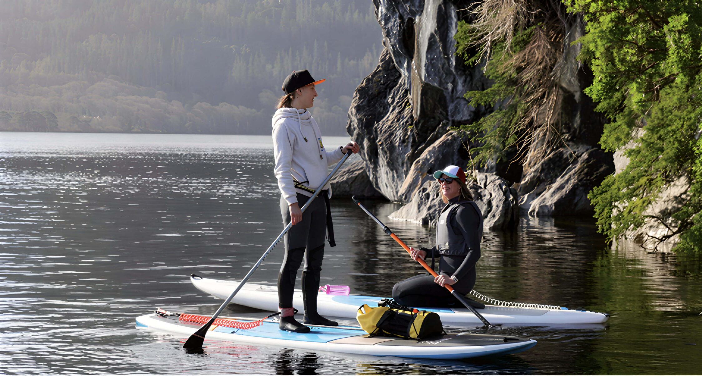 Two people paddleboarding on a serene lake near lush greenery and rocky cliffs.
