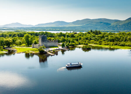 Scenic view of Ross Castle by a lake, surrounded by lush greenery and mountains under a clear sky.