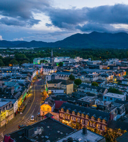 Aerial view of Killarney town at dusk, with glowing streetlights and scenic mountains in the background.