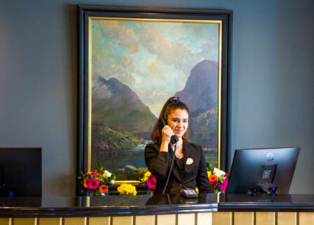 Friendly receptionist at Scotts Hotel Killarney front desk, with a mountain painting and flowers.