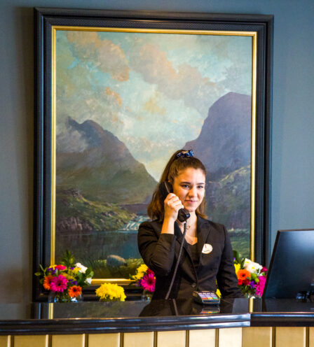 Friendly receptionist at Scotts Hotel Killarney front desk, with a mountain painting and flowers.