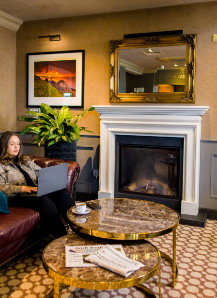 Woman relaxing with a laptop in a cosy hotel lounge by a fireplace at Scotts Hotel Killarney.