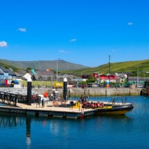 People boarding a yellow boat at Killarney harbour with colourful buildings and hills in the background.