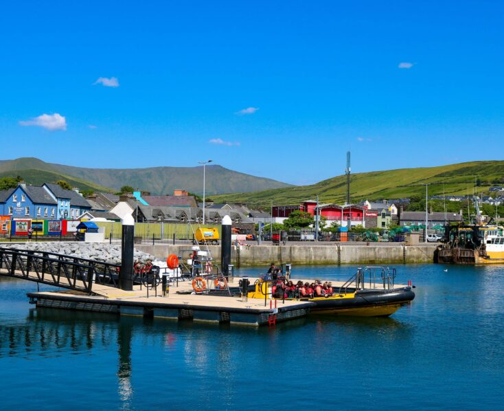 People boarding a yellow boat at Killarney harbour with colourful buildings and hills in the background.
