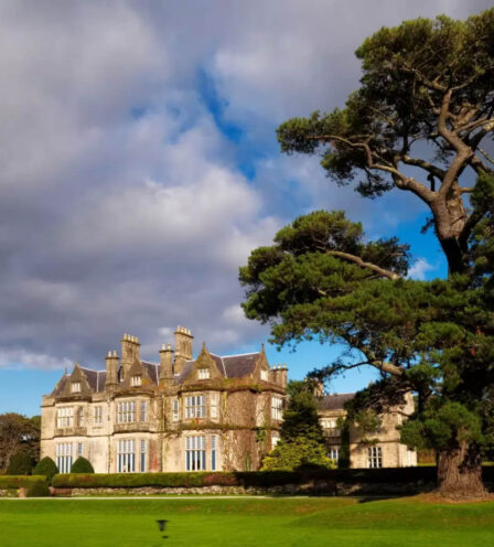 Grand historic hotel with lush garden, large tree, and guests strolling on green lawn in Killarney.