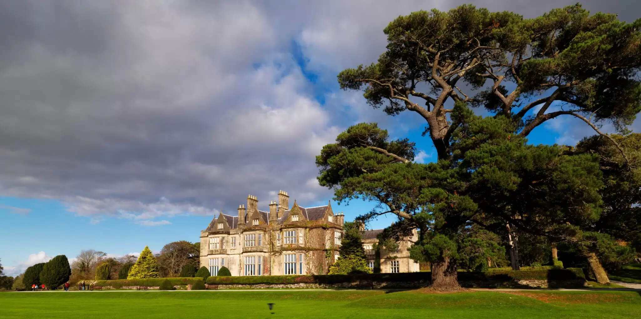 Grand historic hotel with lush garden, large tree, and guests strolling on green lawn in Killarney.