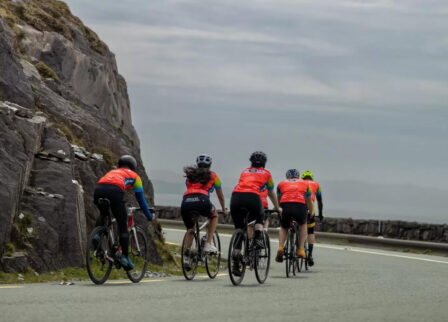 Cyclists in colourful jerseys ride along a scenic road near rocky cliffs and open sky in Killarney.
