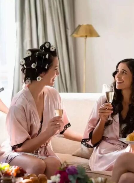 Four women in robes enjoying champagne and pastries in a cosy hotel room.