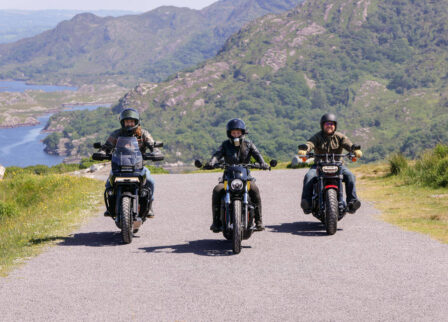 Three motorcyclists ride on a scenic road near lush hills and a tranquil lake in Killarney.