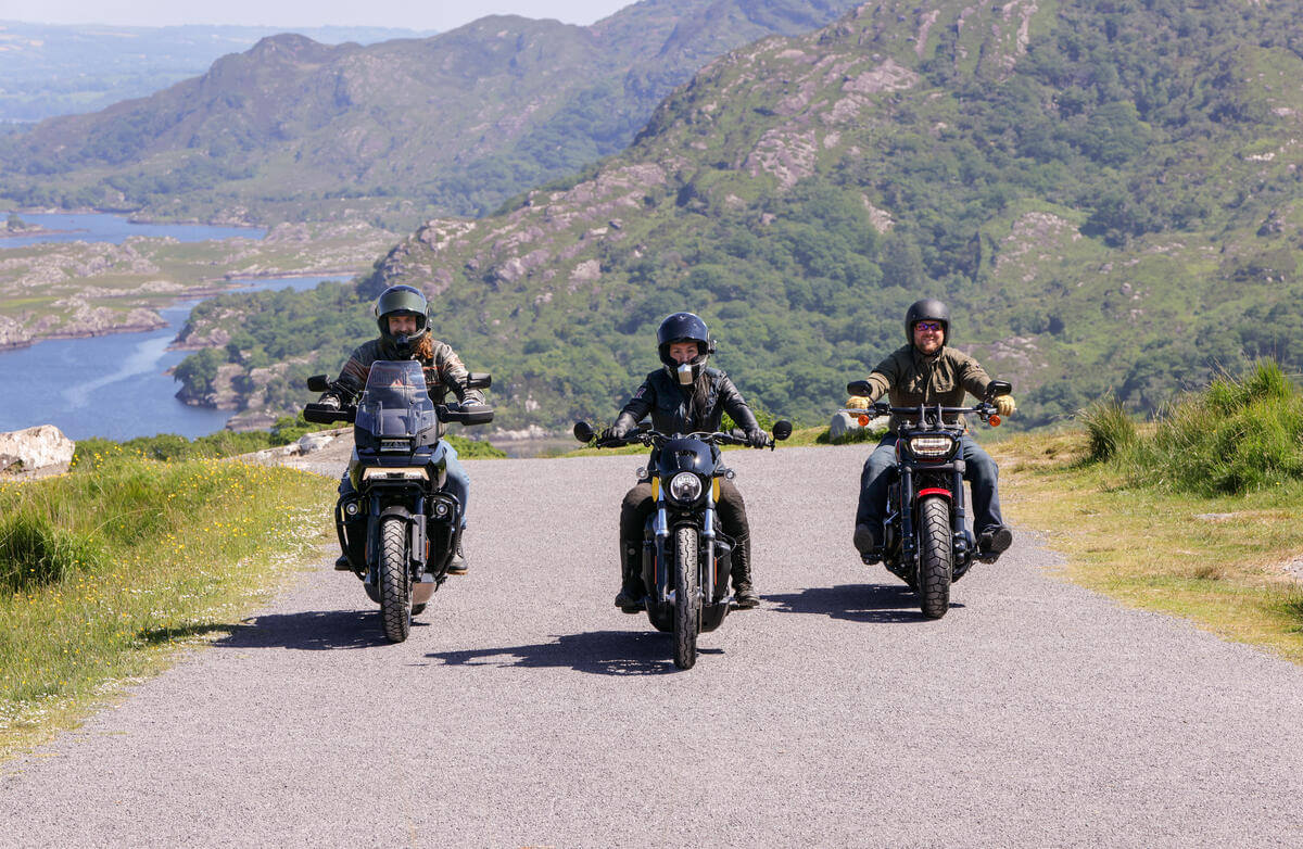 Three motorcyclists ride on a scenic road near lush hills and a tranquil lake in Killarney.