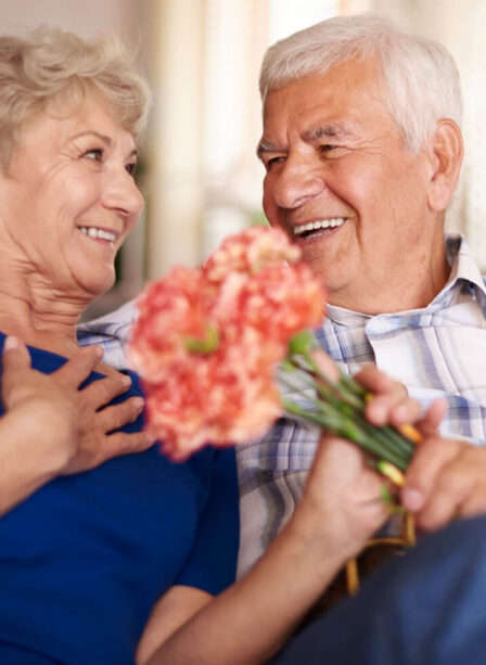 Elderly couple smiling and embracing indoors with flowers.