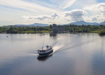 Scenic boat ride on calm lake with lush greenery and distant mountains, near Scotts Hotel Killarney.