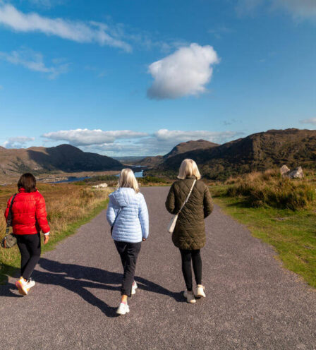 Three women walk on a scenic path with hills and a bright blue sky in Killarney.