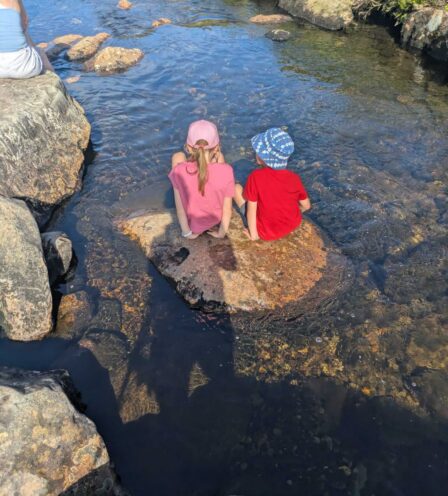 Two children in colourful hats sit on a rock in a clear stream, enjoying a sunny day outdoors.