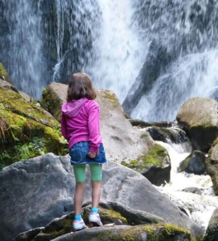 Young girl in pink jacket admires a cascading waterfall amidst mossy rocks.