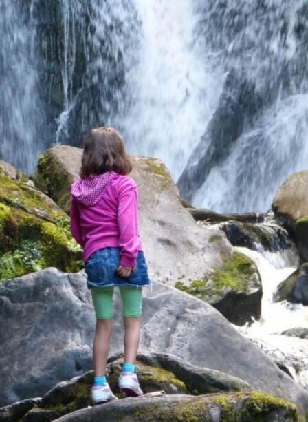 Young girl in pink jacket admires a cascading waterfall amidst mossy rocks.