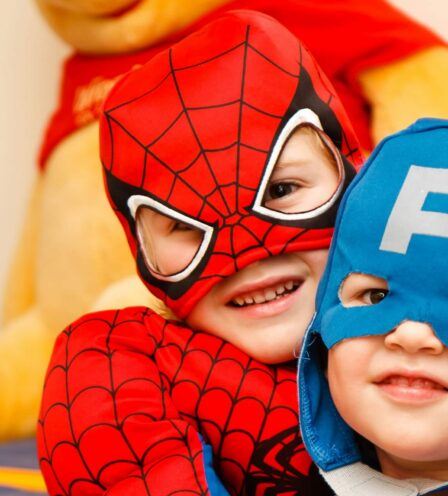 Two children in superhero costumes smiling indoors with plush toy in background.