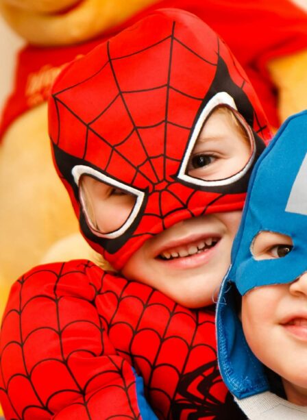 Two children in superhero costumes smiling indoors with plush toy in background.