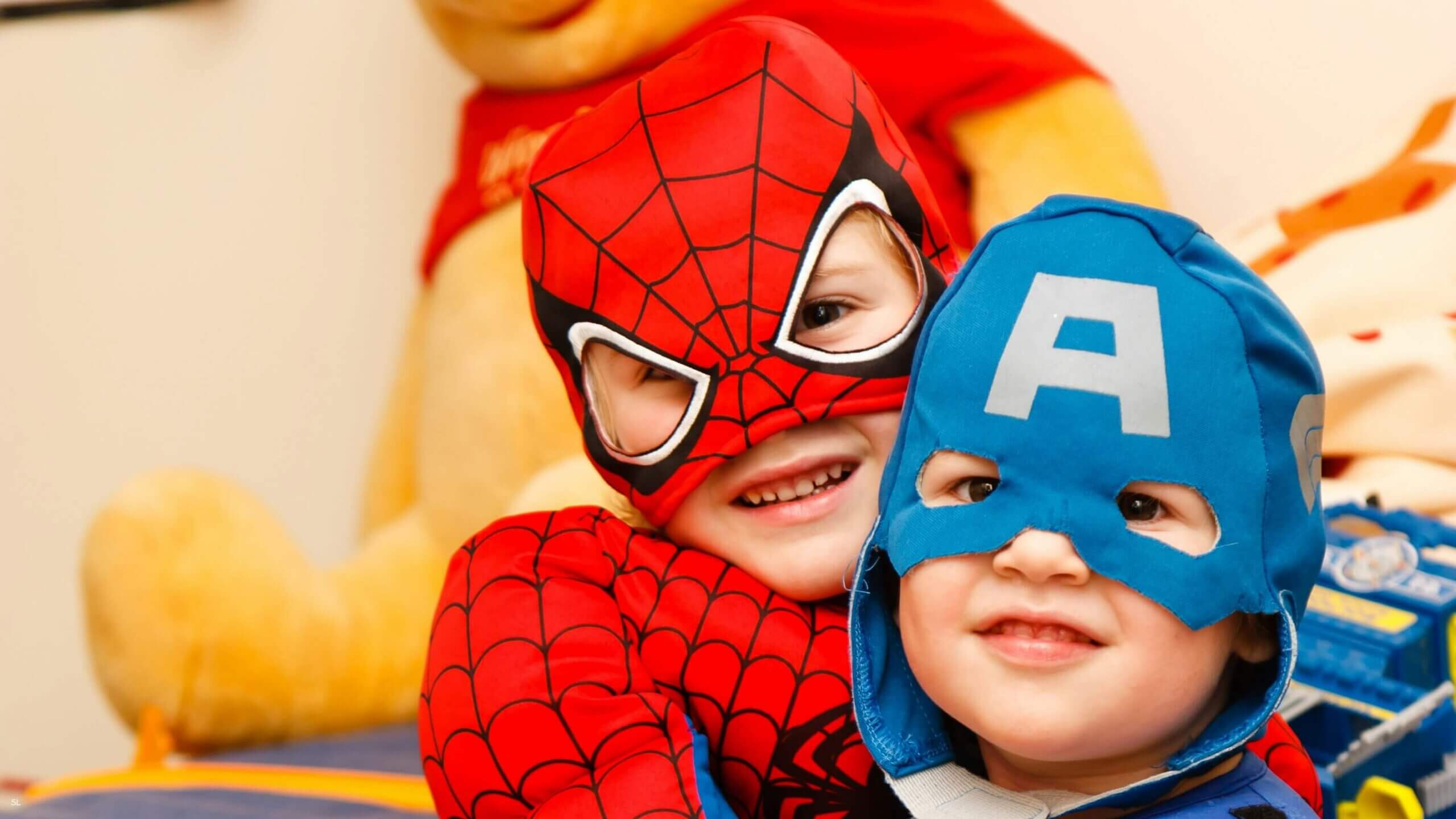 Two children in superhero costumes smiling indoors with plush toy in background.